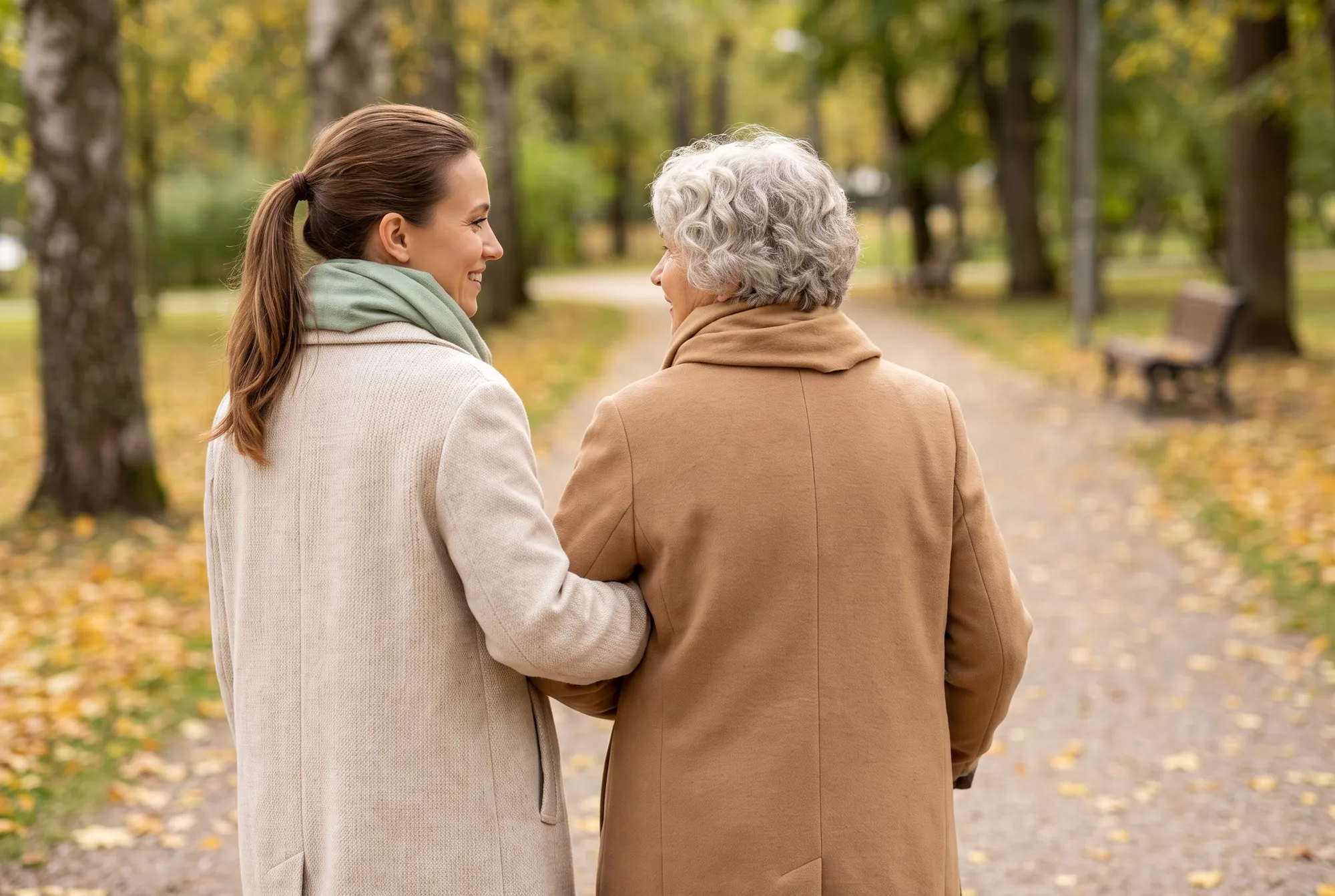 A helper and an older woman walking arm in arm along an autumn avenue